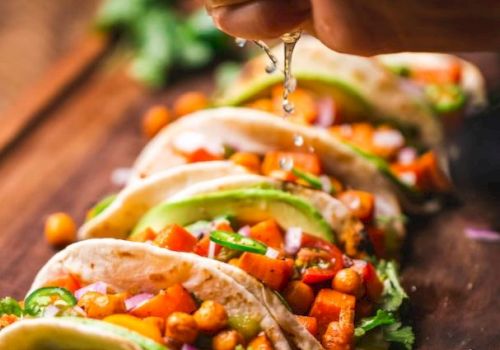 The image shows a hand squeezing a lime over a row of tacos filled with chickpeas, diced vegetables, lettuce, and garnished with cilantro.