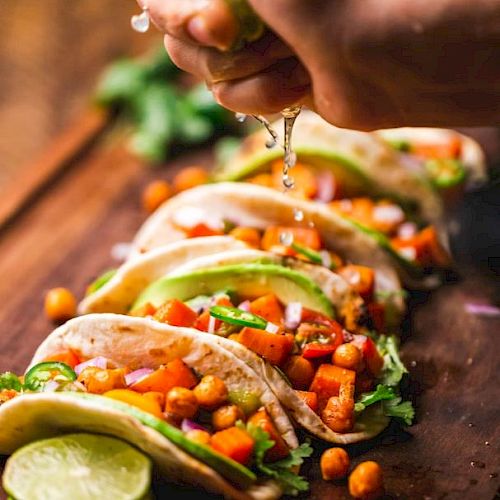 The image shows a hand squeezing a lime over a row of tacos filled with chickpeas, diced vegetables, lettuce, and garnished with cilantro.