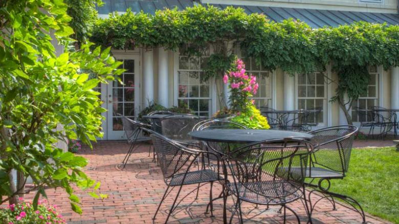 The image shows a patio with several black metal tables and chairs, surrounded by lush greenery and flowering plants, adjacent to a building.